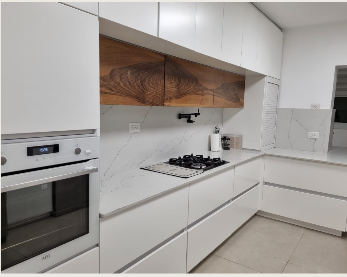 מטבח לבן עם נגרות נקייה, נגיעת עץ טבעי במרכז, משטחי אבן ומראה מינימליסטי מוקפד.